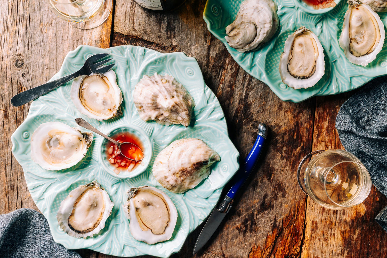 A top-down view of raw oysters served on a decorative mint green ceramic platter with a small dipping sauce bowl, set on a rustic wooden table with a knife, fork, and wine glass nearby.